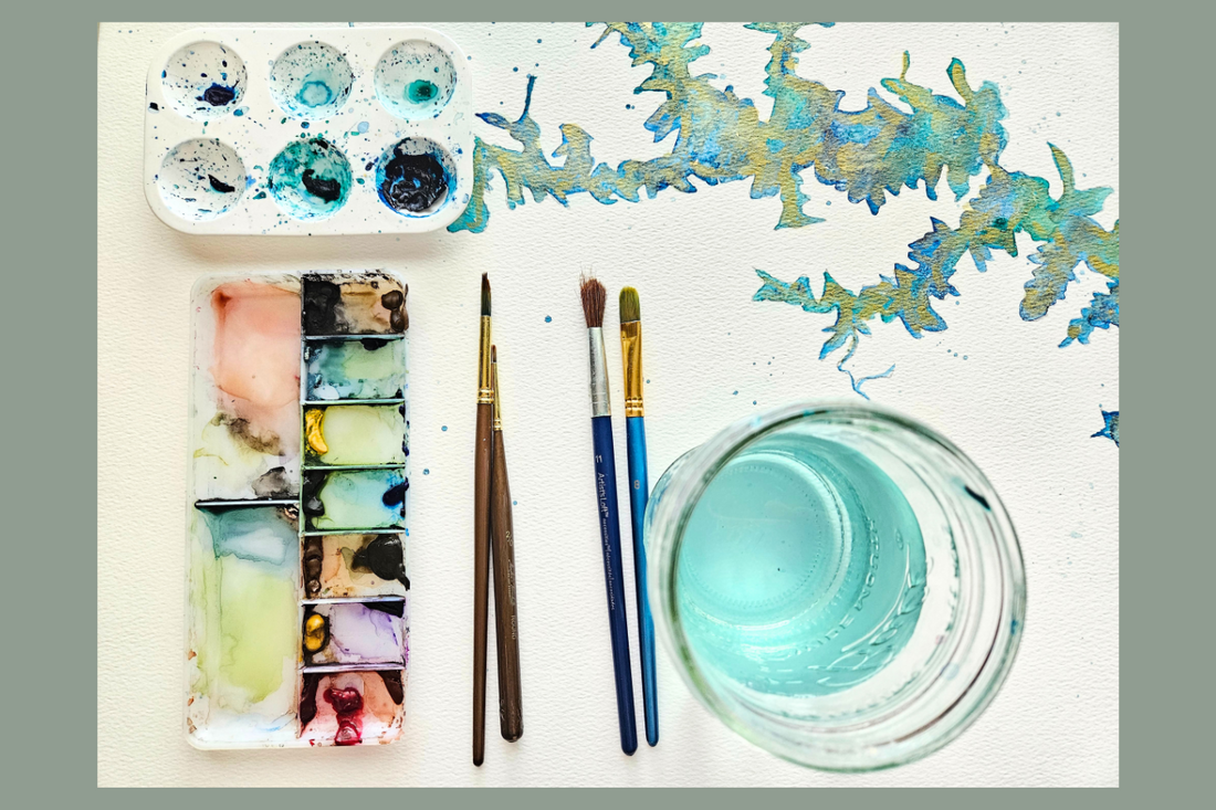 watercolor paints and supplies. palette, paints and brushes on a desk with a jar of water.