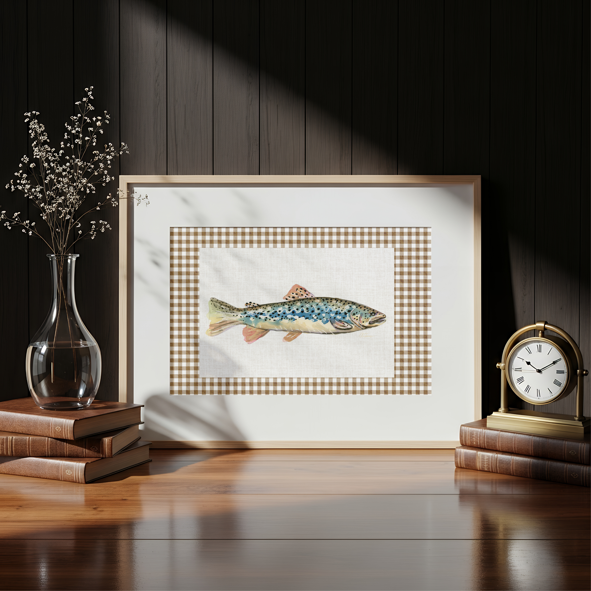 A framed art print featuring a detailed illustration of a blue and orange trout, styled on a wooden table with books, a glass vase, and a clock against a dark wood-paneled wall.